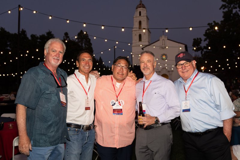 Five members of the class 1984 smile under string lights, the Chapel, and a twilight sky