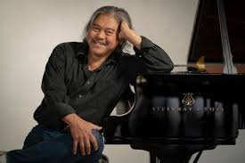 Lino Rivera leaning against his piano with one hand, smiling towards camera