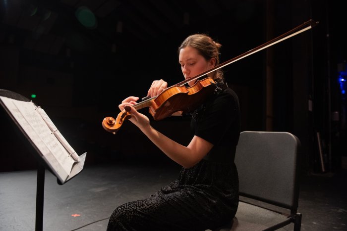 Young woman playing violin with black background and a music stand with sheet music in front of her.